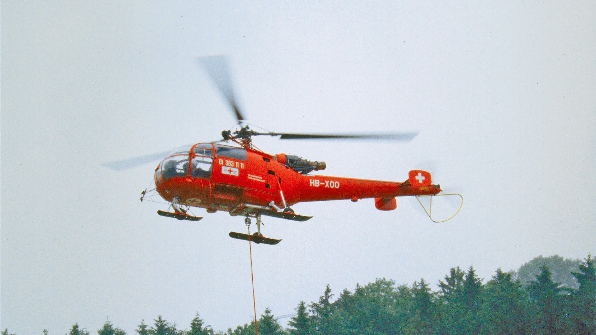 Elicottero rosso Alouette III in volo sopra una foresta.
