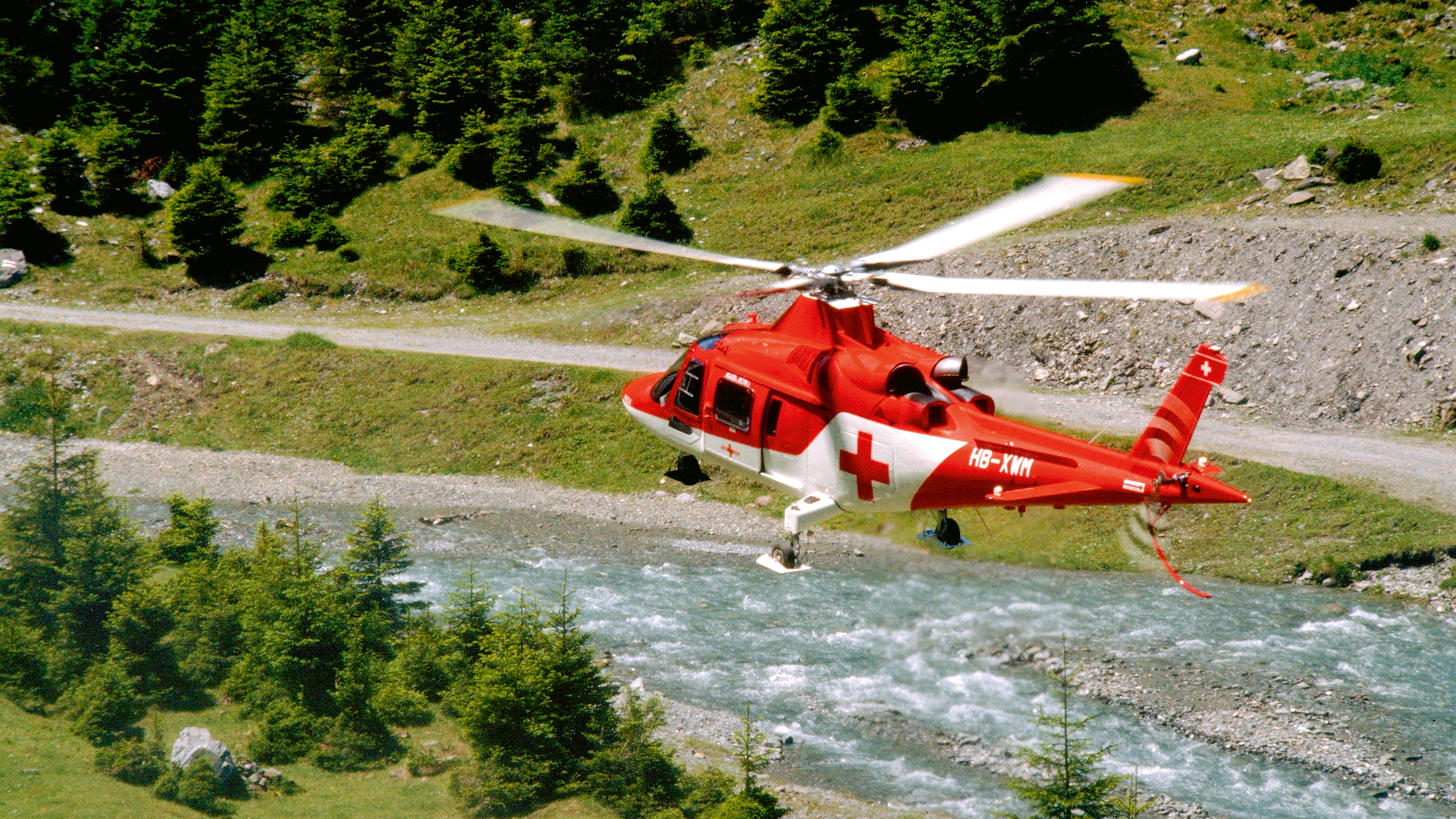Elicottero rosso Agusta A109K2 sopra un fiume nelle Alpi.