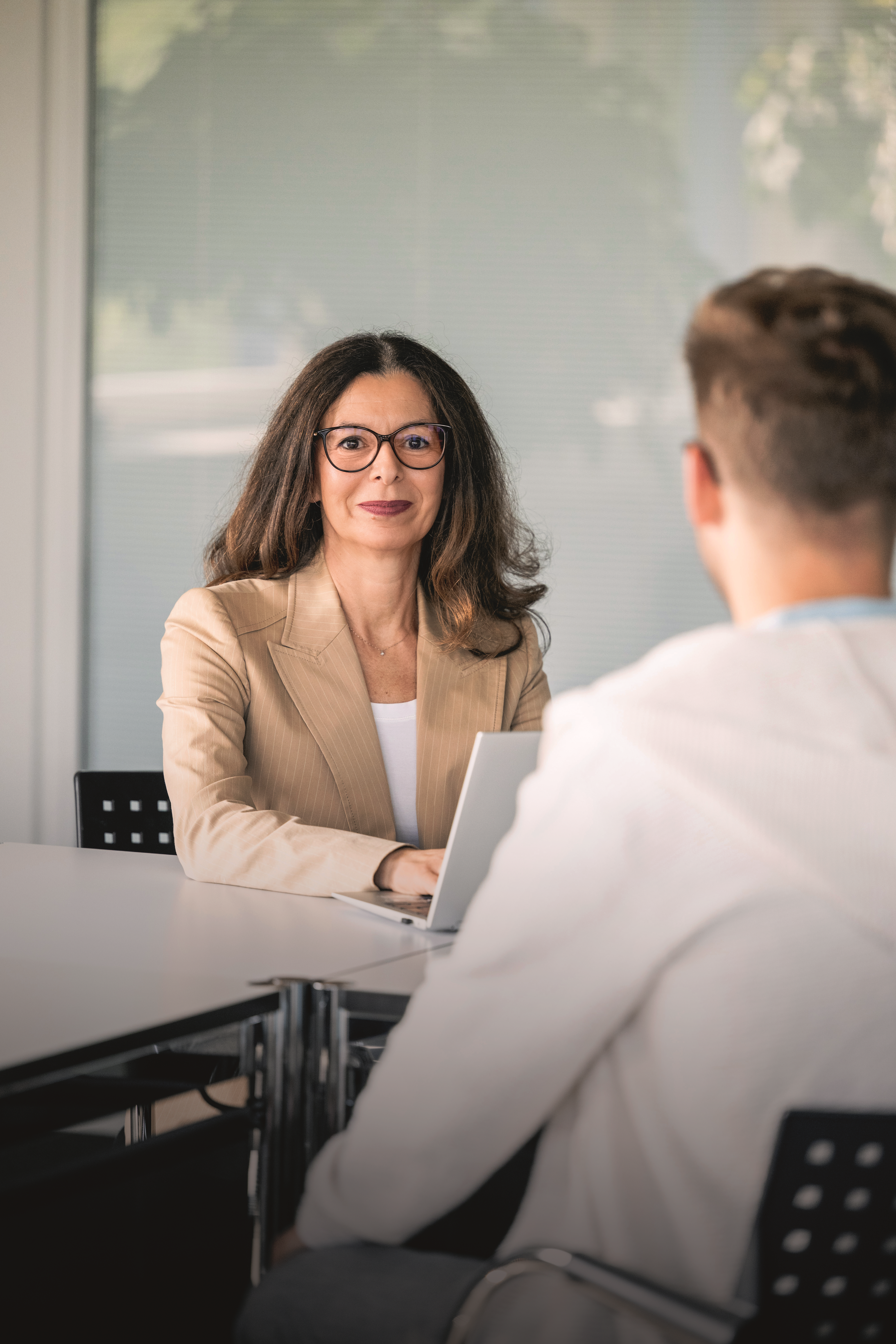 Une femme vêtue de manière professionnelle, portant des lunettes et un blazer beige, est assise à une table. Elle regarde directement l'appareil photo, tandis qu'un jeune homme en pull à capuche blanc est assis au premier plan, dos à la caméra. Un ordinateur portable ouvert est posé sur la table.