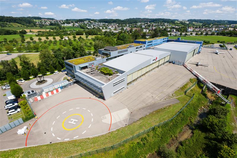 Eine Luftaufnahme des Rega-Centers am Flughafen Zürich-Kloten. Der moderne, mehrteilige Gebäudekomplex mit weissen Hangars und begrüntem Dach ist umgeben von einer Landebahn mit einem deutlich sichtbaren Helikopterlandeplatz. Im Hintergrund ist eine grüne Landschaft mit einem Dorf zu sehen.